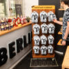 Ein Kunde in einem Laden mit dem Schild "HANDMADE IN BERLIN" nimmt ein Paar schwarz-weiße Flip-Flops von einem Holzständer. Auf dem Boden liegt eine Fußmatte mit der Aufschrift "CHECK YOUR SIZE", auf der Schuhgrößen eingezeichnet sind.