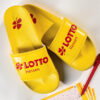 Ein Paar gelbe Badeschlappen mit dem roten Lotto-Hessen-Logo liegt neben einem ausgefüllten Lottoschein und einem roten Stift.