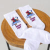 Ein Paar weiße Socken mit dem Logo der "Young Roosters" und dem personalisierten Namen "HEIKE #07" liegt auf einem Holzhocker.
