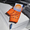 Ein Paar orangefarbene Socken mit blauen Punkten und dem Schriftzug "Emma" liegt auf einer Matratze. Daneben ist eine Karte mit dem Text "Awaken Your Best" zu sehen.