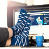 Eine Person sitzt am Schreibtisch mit den Füßen auf dem Tisch und trägt dunkelblaue Socken mit weißen Streifen. Im Hintergrund ist ein Computermonitor mit der Startseite von "IONOS" zu sehen.