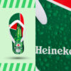 Ein einzelner Flip-Flop mit weißem Riemen, der auf einem hellgrünen Hintergrund schwebt. Die Sohle ist mit einer grünen Heineken-Bierflasche bedruckt. Im oberen rechten Bildausschnitt ist eine vergrößerte Ansicht des Heineken-Logos und von Wassertropfen auf einer grünen Oberfläche zu sehen.