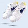 Par de zapatillas blancas de cuero sintético mostradas en ángulo. Presentan un logotipo lateral en forma de "W" azul oscuro y un detalle amarillo vibrante en el talón. Cordones blancos y diseño limpio.