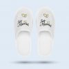 A white pair of slippers made of waffle piqué fabric with “Just Married” embroidery and two gold rings.